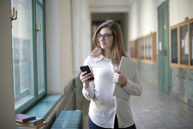 a woman standing next to a window holding a cell phone, in school hallway, nerdy, trending photo, holding notebook