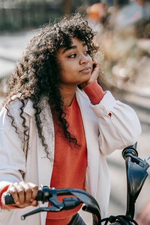 a woman sitting on a bike talking on a cell phone, trending on pexels, happening, black curly hair, pensive, wearing jacket, wearing a white sweater