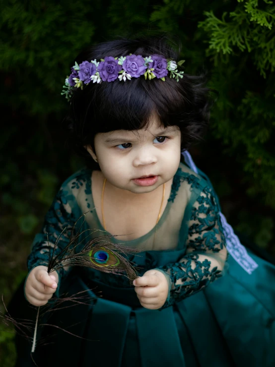 a little girl in a green dress holding a peacock feather, a portrait, pexels contest winner, dark purple crown, flowers in her dark hair, toddler, & her expression is solemn