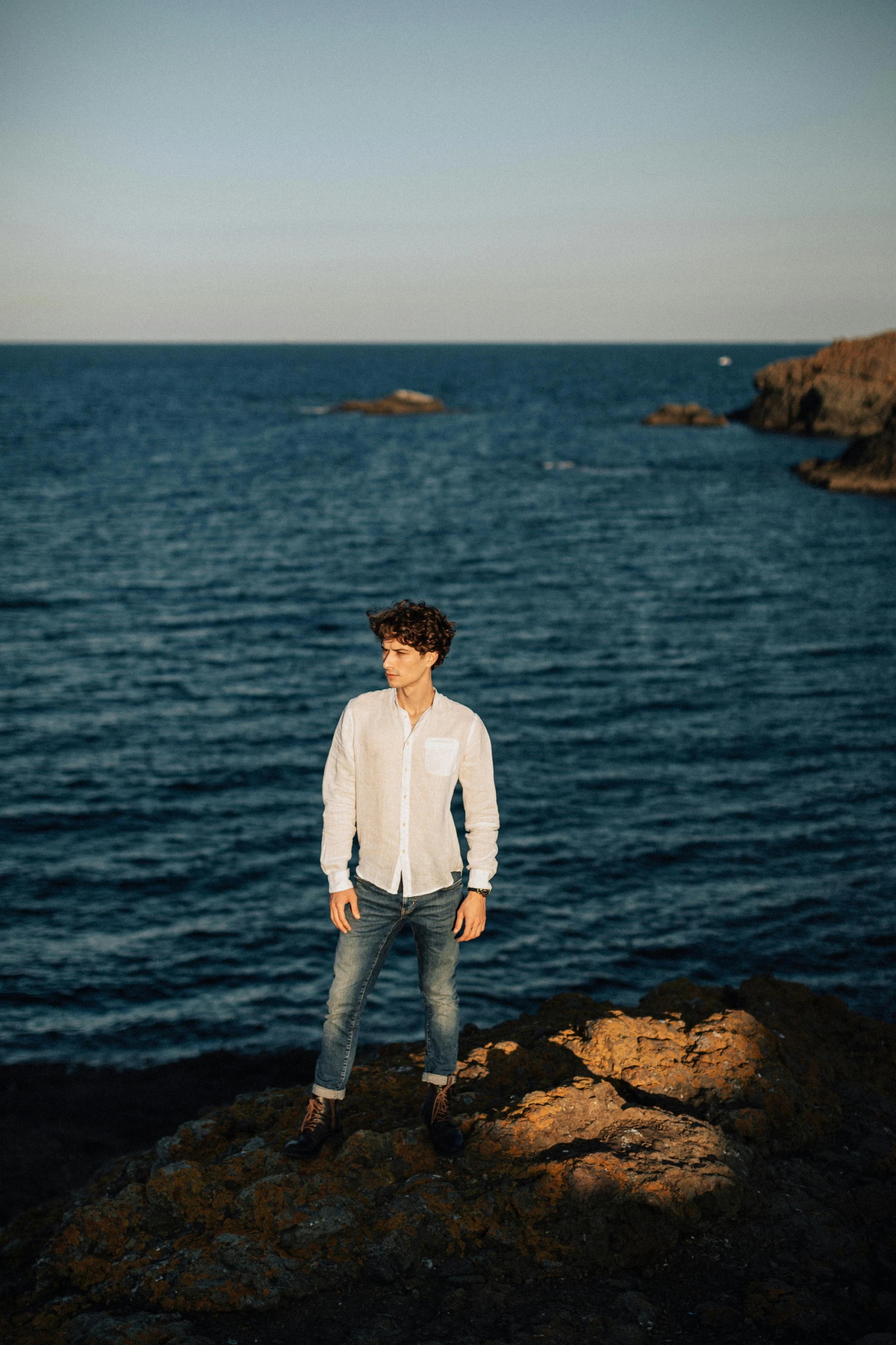 a man standing on top of a rock next to the ocean, an album cover, by Adam Dario Keel, pexels contest winner, renaissance, white shirt and jeans, male ulzzang, androgynous male, riccardo scamarcio