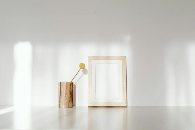 a picture frame sitting on top of a table next to a vase, pexels contest winner, light and space, white and yellow scheme, background image, cream white background, miscellaneous objects