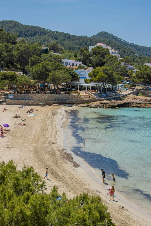 a group of people standing on top of a sandy beach, ibiza, white buildings with red roofs, kids playing at the beach, sparkling cove