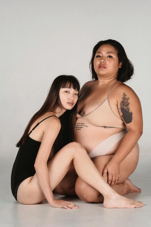 a couple of women sitting next to each other, an album cover, inspired by Wang Duo, unsplash, realism, her belly is fat and round, half asian, bodysuit, nonbinary model