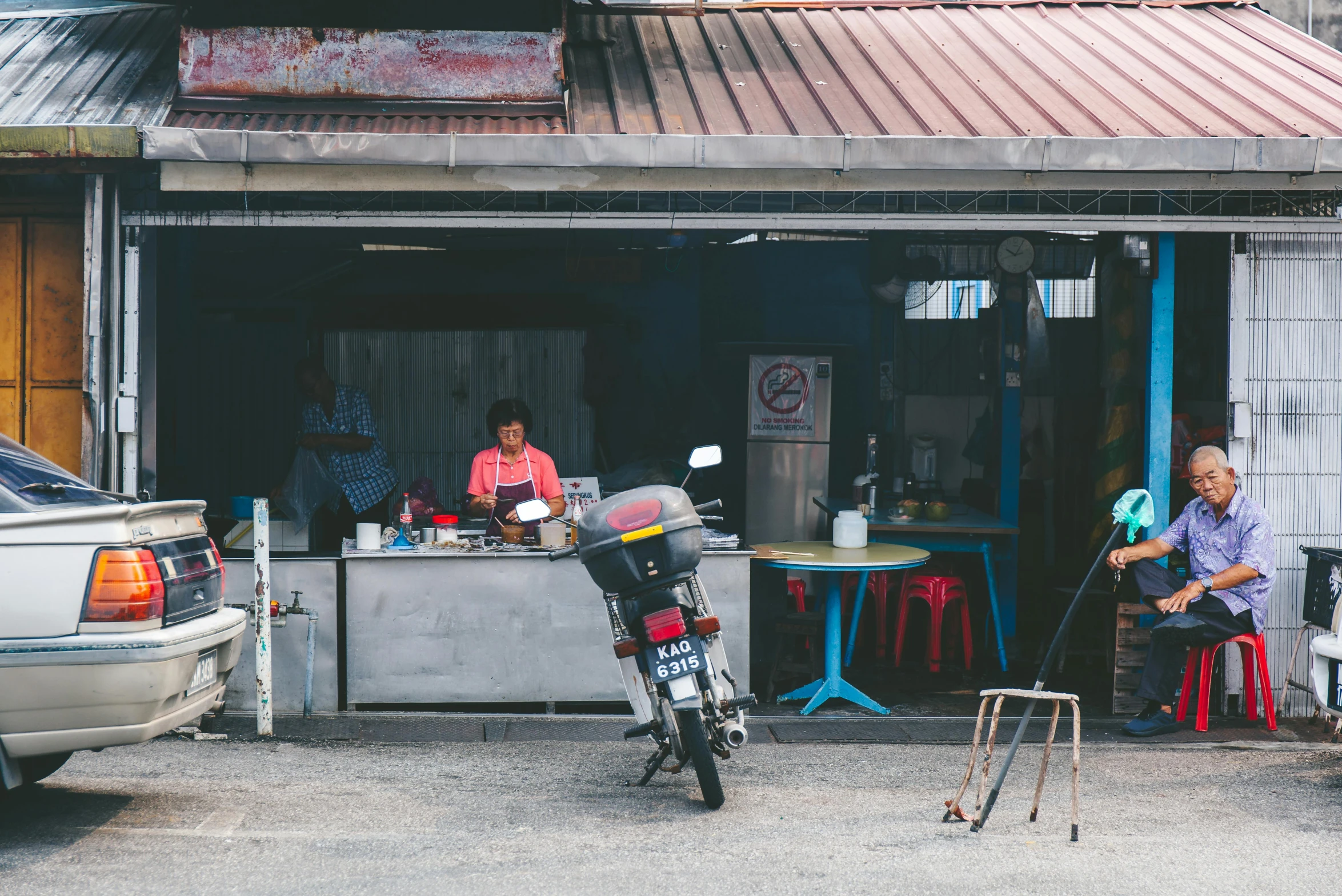 a couple of people sitting at a table in front of a building, pexels contest winner, malaysian, in front of a garage, merchant stands, moped