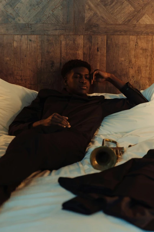 a man laying on top of a bed next to a lamp, an album cover, pexels contest winner, trumpet, lance reddick, moody aesthetic, gold suit