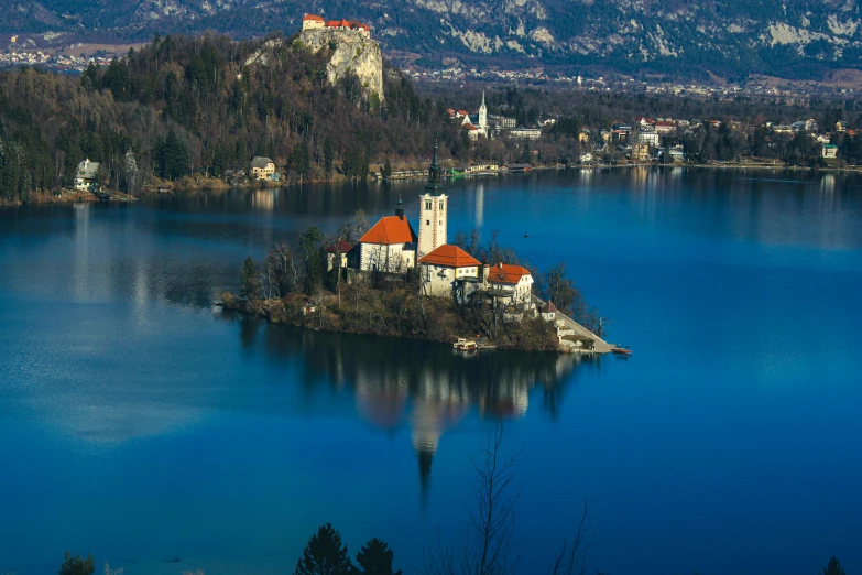 a small island in the middle of a lake, by Matija Jama, pexels contest winner, art nouveau, majestic spires, blue waters, mountains and lakes, white buildings with red roofs