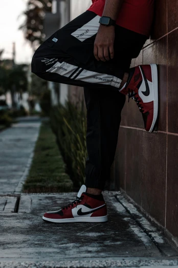 a man leaning against a wall wearing a red shirt and black pants, by Niko Henrichon, trending on pexels, air jordan 1 high, maroon and white, metallic reflective, standing in an arena