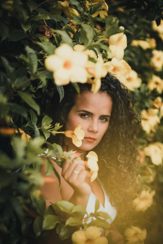 a woman standing in front of a bush of flowers, inspired by Elsa Bleda, pexels contest winner, wavy hair yellow theme, hispanic, staring at you, medium format