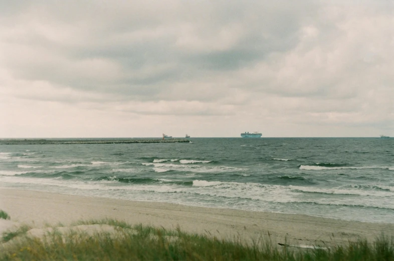 a man standing on top of a sandy beach next to the ocean, a colorized photo, inspired by Elsa Bleda, realism, ships in the harbor, overcast gray skies, shipfleet on the horizon, vsco film grain