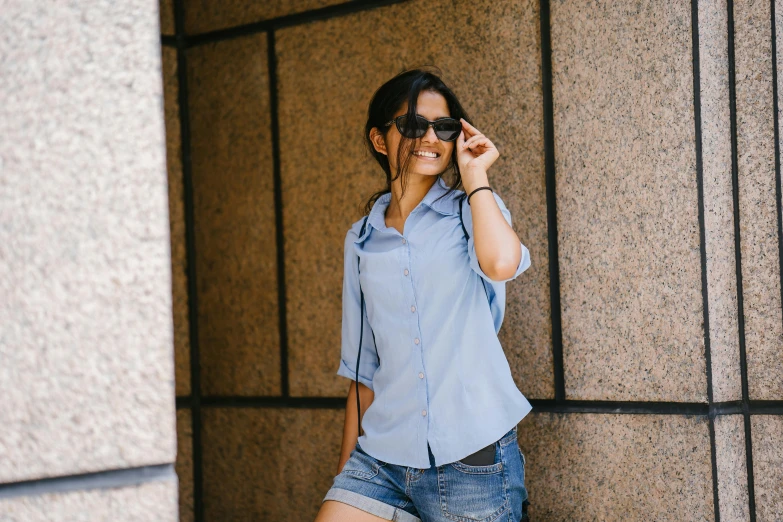 a woman leaning against a wall talking on a cell phone, pexels contest winner, wearing blue sunglasses, wearing a light blue shirt, avatar image, off the shoulder shirt