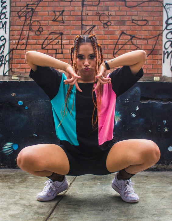 a woman squatting in front of a wall with graffiti on it, an album cover, by Amelia Peláez, unsplash, wearing shorts and t shirt, lesbians, classic dancer striking a pose, colorful pigtail