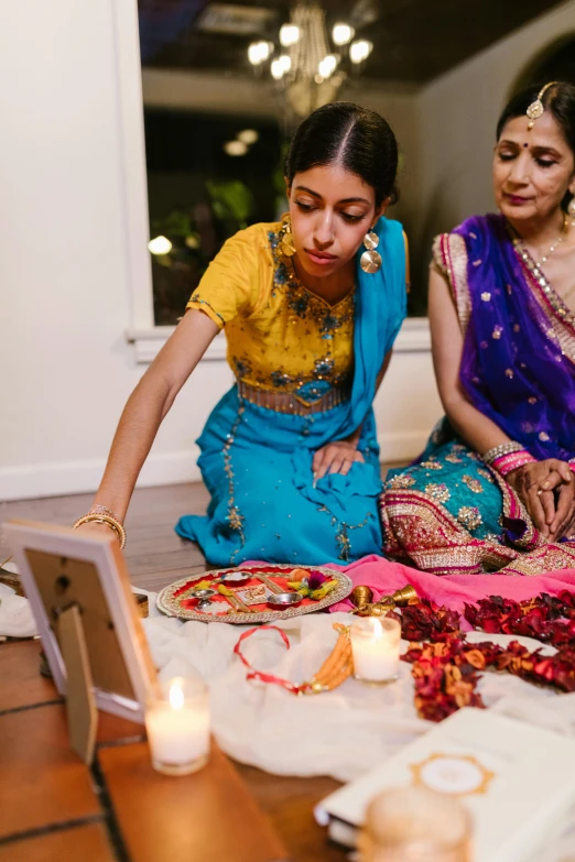 a couple of women sitting on top of a floor, reddit, hurufiyya, festive colors, candles in foreground, bollywood, square