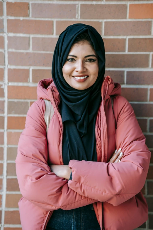 a woman standing in front of a brick wall, inspired by Maryam Hashemi, pexels contest winner, hurufiyya, wearing jacket, warm friendly expression, student, asian human