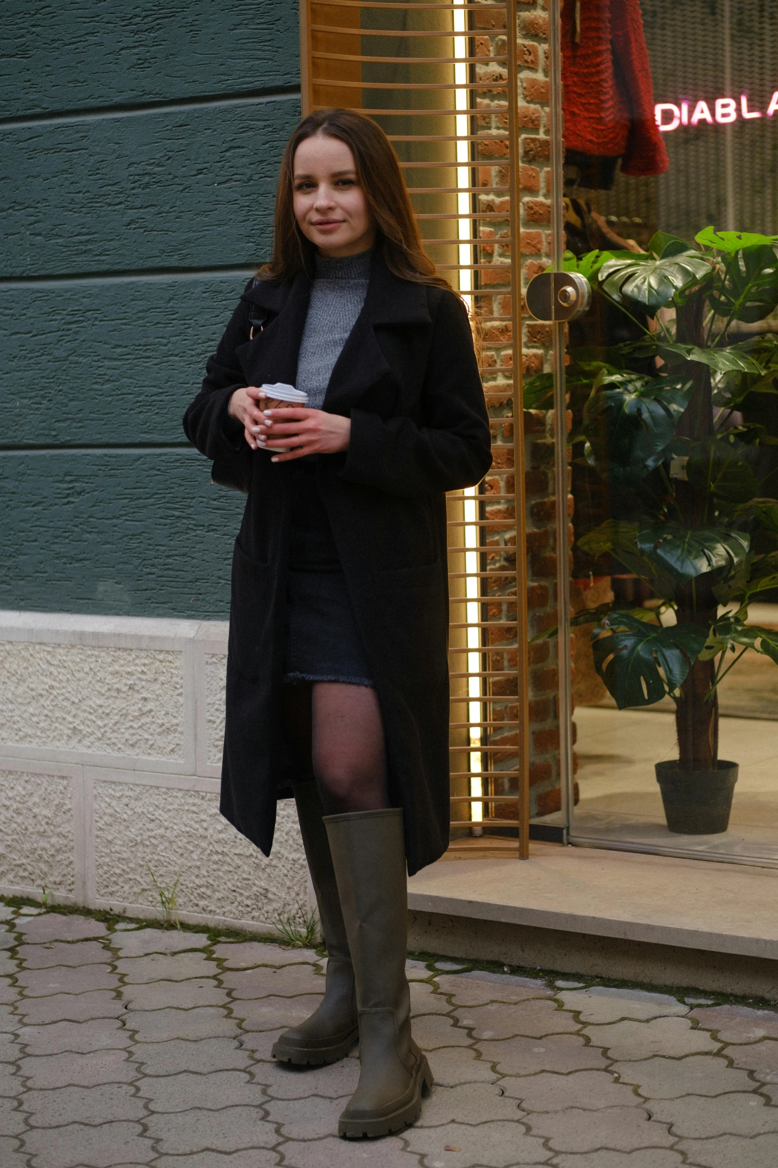 a woman standing in front of a building holding a cup of coffee, lady in black coat and pantyhose, model エリサヘス s from acquamodels, leather boots, cold as ice! 🧊