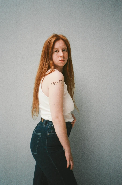 a woman standing in front of a gray wall, an album cover, by Carey Morris, trending on pexels, hyperrealism, long ginger hair, ( ( ( wearing jeans ) ) ), patricia piccinini, queer woman