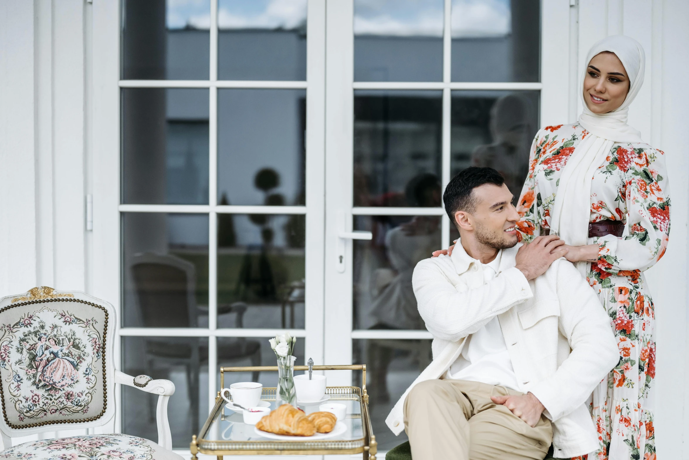 a man sitting on a chair next to a woman, by Emma Andijewska, pexels contest winner, romanticism, white hijab, background image, at the terrace, family friendly