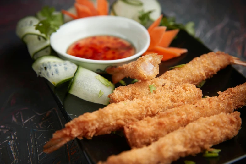 a black plate topped with shrimp sticks next to a bowl of dipping sauce, inspired by Kōno Michisei, unsplash, hurufiyya, square, vietnam, shiny crisp finish, children's