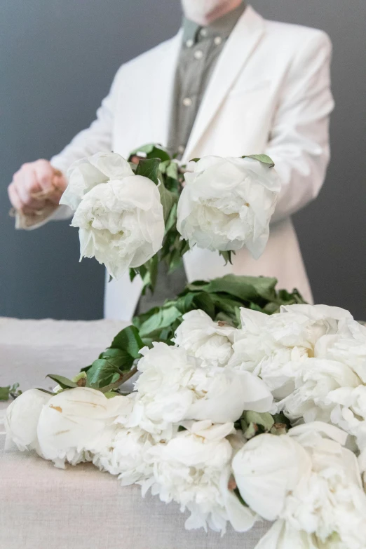 a man standing in front of a bunch of white flowers, a marble sculpture, inspired by François Boquet, romanticism, white lab coat, hands on counter, peony flower, funeral