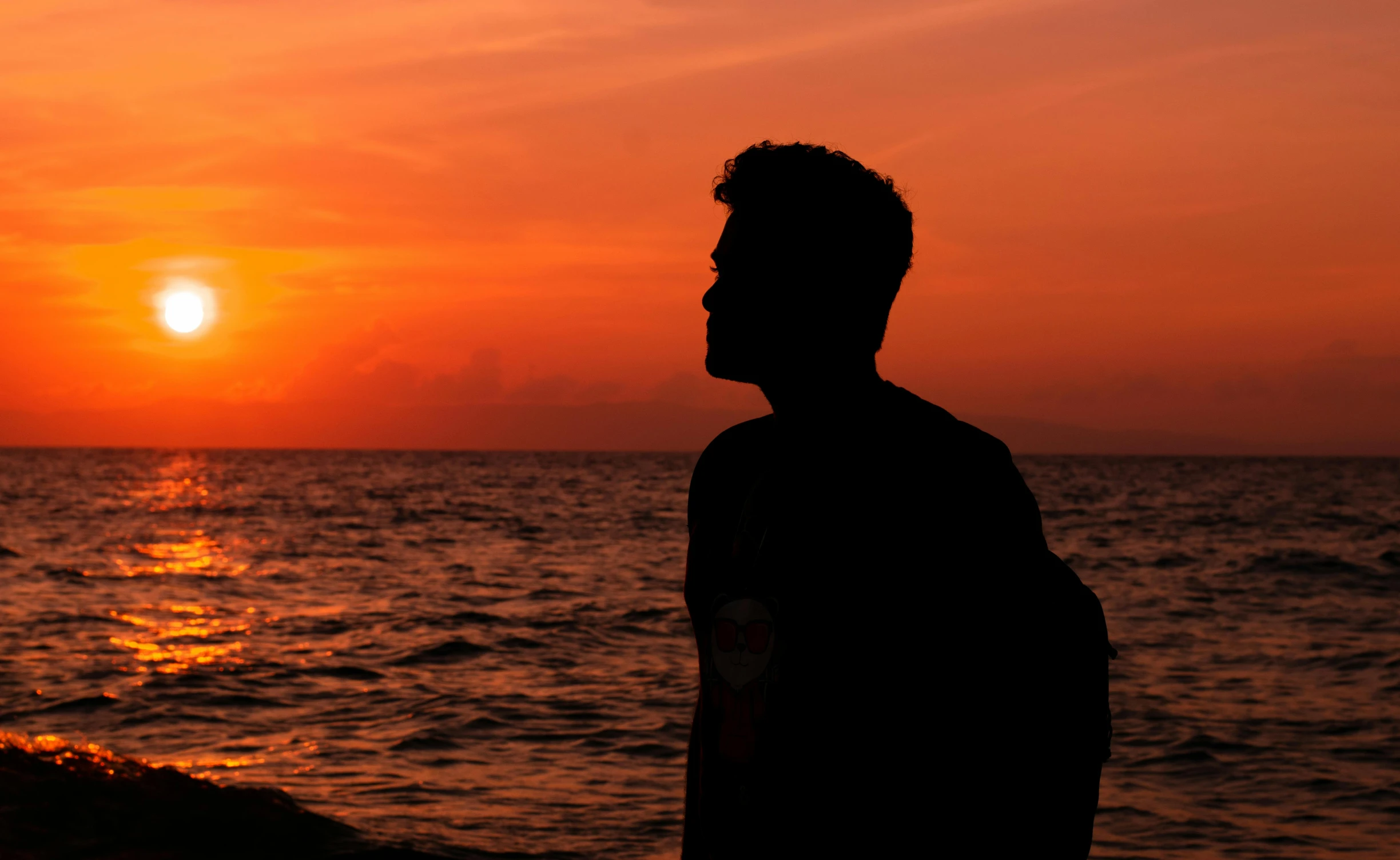 a man standing on top of a beach next to the ocean, by Robbie Trevino, pexels contest winner, romanticism, backlit face, orange and red sky, profile image, outlined silhouettes