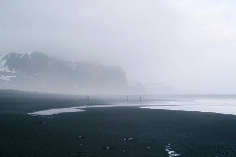 a man standing on top of a beach next to the ocean, by Andrew Geddes, pexels contest winner, romanticism, uneven dense fog, a group of people, iceland, mermaids in distance