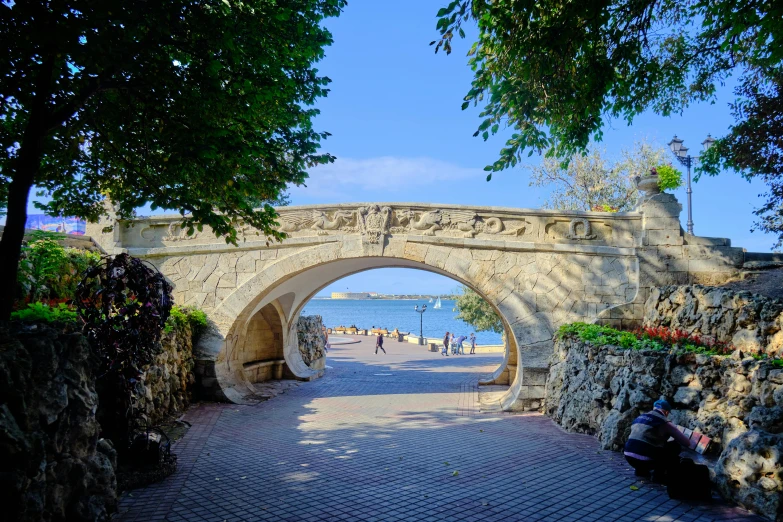 a person sitting on a bench under a bridge, by Tom Wänerstrand, pexels contest winner, art nouveau, summer street near a beach, stone grotto in the center, manly, people walking around