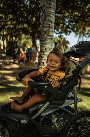 a small child sitting in a stroller in a park, a portrait, by Drew Tucker, pexels contest winner, waikiki beach, battle ready, sturdy body, complex background