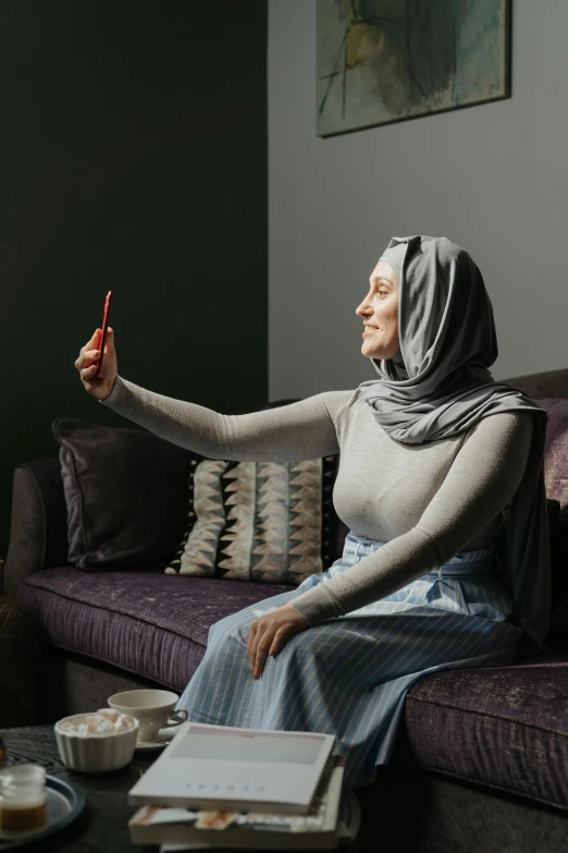 a woman sitting on a couch taking a selfie, inspired by Maryam Hashemi, hurufiyya, tall shot, grey, modest, interacting and posing