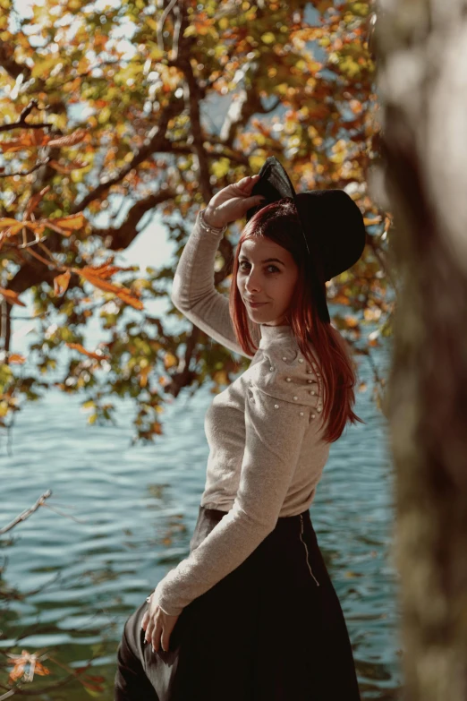 a woman standing next to a tree near a body of water, wearing a tophat, copper hair, 🍂 cute, pose 4 of 1 6