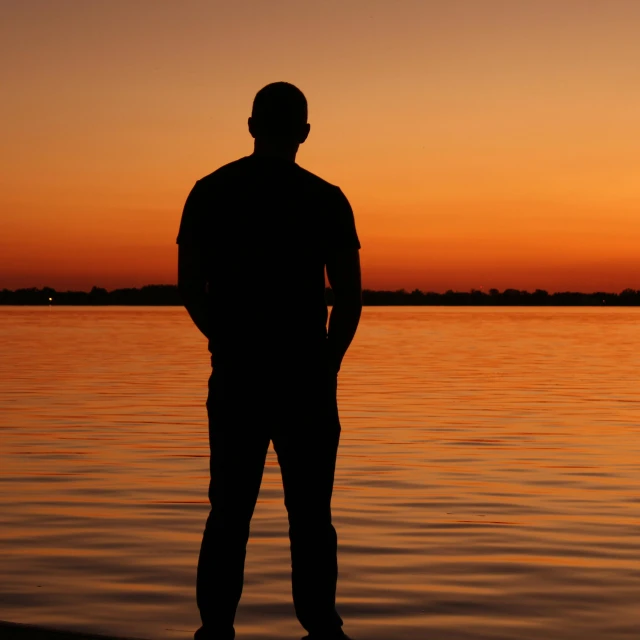 a man standing in front of a body of water at sunset, in the sunset