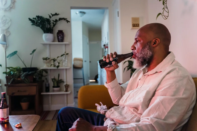 a man sitting on a couch drinking from a bottle, by Carey Morris, pexels contest winner, happening, old gigachad with grey beard, black man, holding a small vape, in a living room