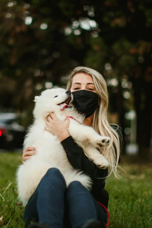 a woman sitting in the grass holding a white dog, wearing all black mempo mask, lifestyle, fluffy, blond