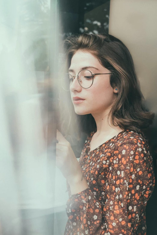 a woman wearing glasses looking out a window, a picture, inspired by Elsa Bleda, trending on pexels, renaissance, girl with brown hair, square rimmed glasses, attractive photo, lorde