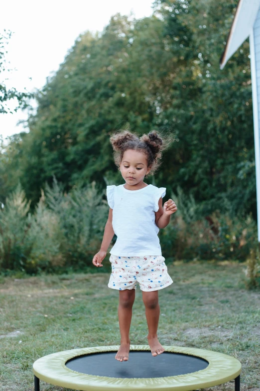 a little girl that is standing on a trampol, by Lucia Peka, wearing shorts and t shirt, nature outside, midsommar color theme, light skinned african young girl