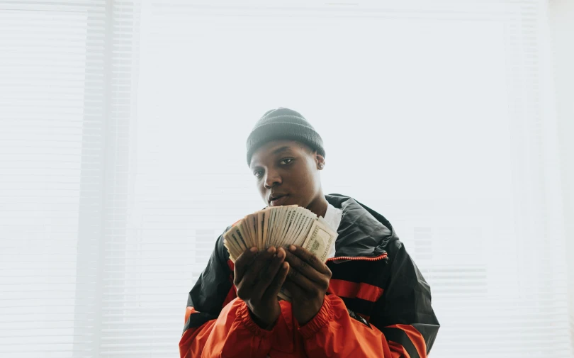 a man holding a stack of money in front of a window, an album cover, pexels contest winner, black teenage boy, hq 4k phone wallpaper, robbery, looking straight