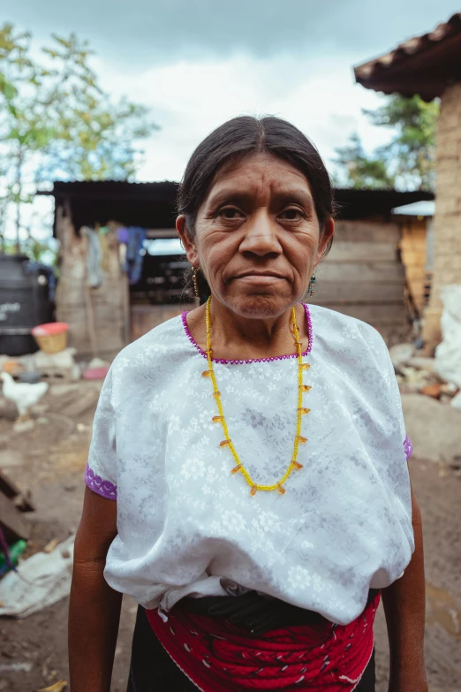 a woman standing in front of a shack, by Alejandro Obregón, portrait image, elder, looking serious, wearing a dress made of beads