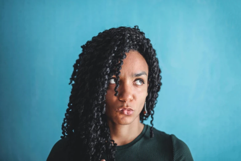 a woman with curly hair standing in front of a blue wall, an album cover, by Lily Delissa Joseph, pexels contest winner, antipodeans, long black braided hair, angry looking at camera, high angle closeup portrait, ashteroth