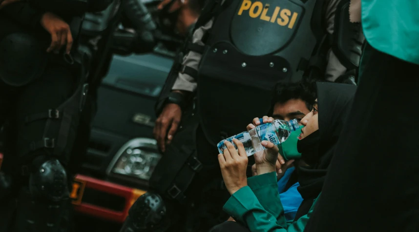 a man sitting on the ground with a bottle of water in his hand, pexels contest winner, realism, police officers under heavy fire, jakarta, girl drinks monster energy, riot shields