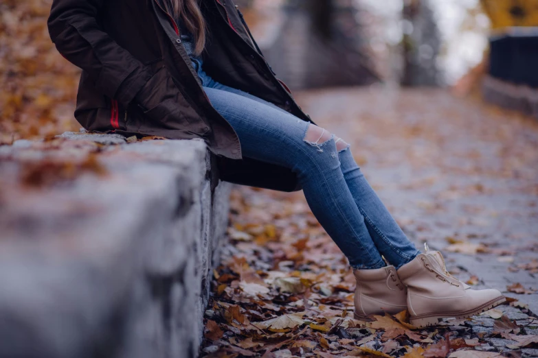 a woman sitting on a ledge in a park, trending on pexels, jeans and boots, leafs, brown jacket, realistic »