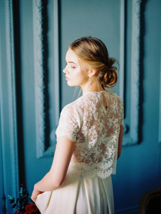 a woman standing in front of a blue wall, by Sara Saftleven, dressed in a frilly ((lace)), girl with messy bun hairstyle, open back dress, beautiful girl in an empty room