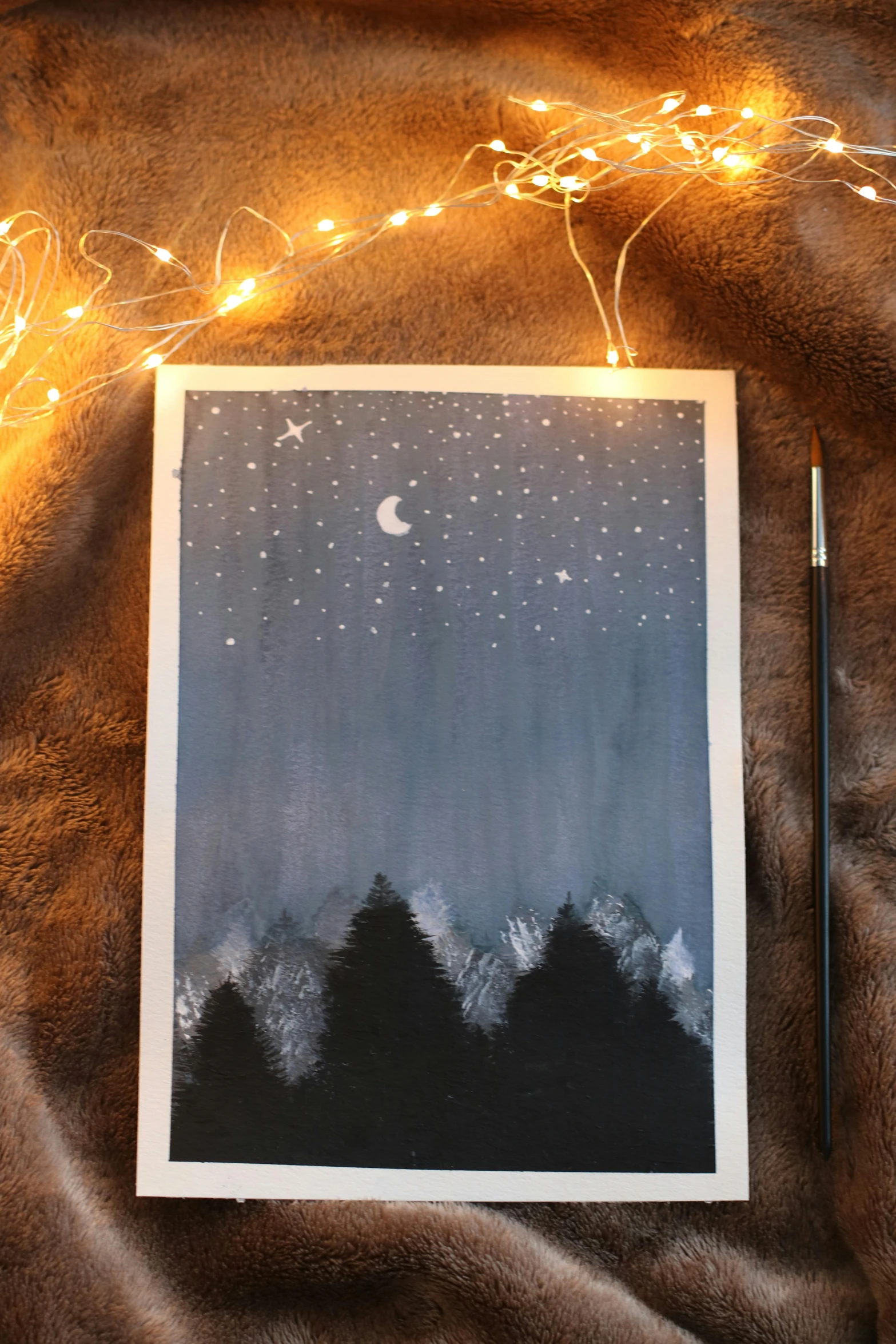 a card sitting on top of a blanket next to a string of lights, an acrylic painting, an image of a moonlit forest, black sky with stars, pastel style painting, ☁🌪🌙👩🏾