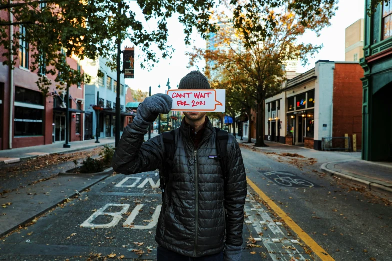 a man holding a sign in front of his face, by Carey Morris, unsplash, street art, fall season, bentonville arkansas, thumbnail, wintermute