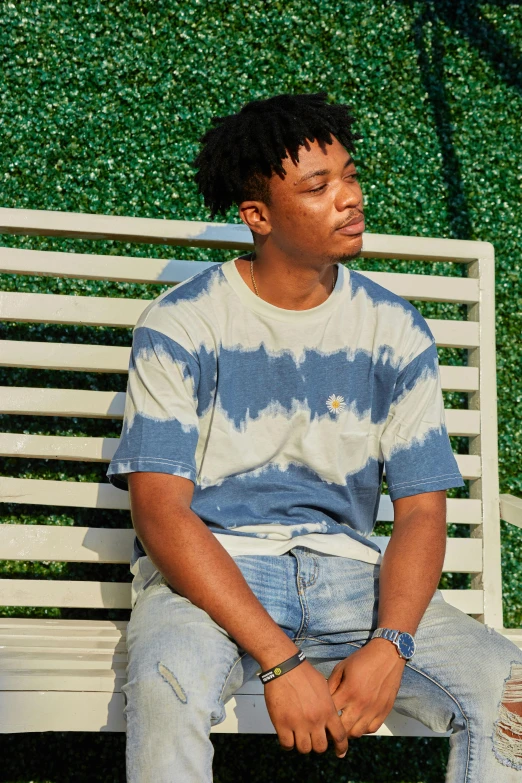a man sitting on top of a white bench, wearing a tie-dye t-shirt, yzy gap, next to a plant, washed out background