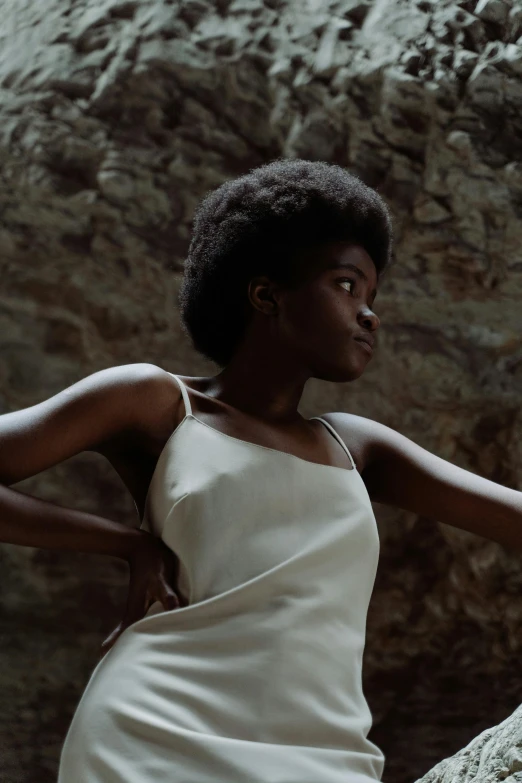 a woman in a white dress leaning against a rock, inspired by Gordon Parks, afrofuturism, natural hair, perfectly lit. movie still, wearing a camisole, ignant