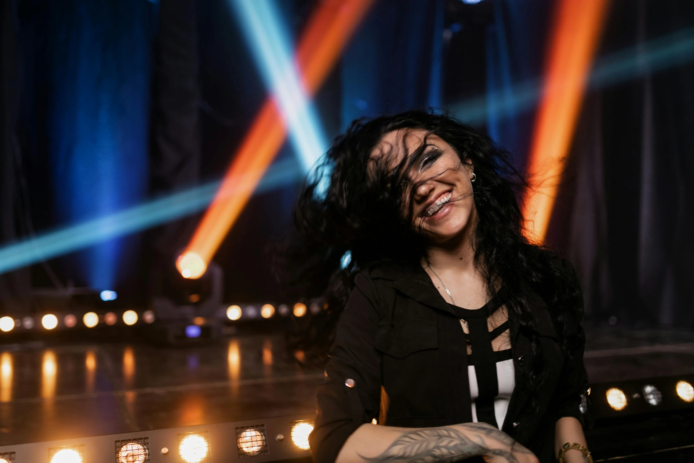 a woman that is standing in front of some lights, with tattoos, woman with black hair, laughing, concert photo