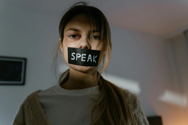a woman with a tape over her mouth that says speak, by Emma Andijewska, trending on pexels, woman holding sign, from the waist up, light stubble, covid-19 as a human