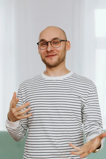 a man standing in front of a green couch, a character portrait, inspired by Alexander Kanoldt, pexels contest winner, wearing stripe shirt, bald, jewish young man with glasses, waving at the camera