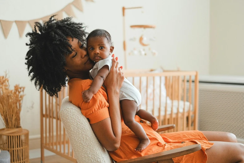 a woman sitting in a rocking chair holding a baby, by Lily Delissa Joseph, pexels contest winner, wearing an orange jumpsuit, young black woman, nursery poster, hugging