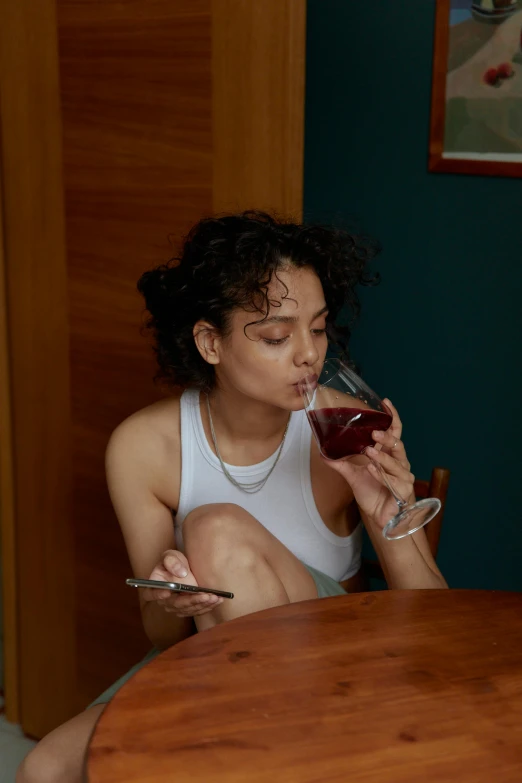 a woman sitting at a table with a glass of wine, inspired by Nan Goldin, pexels, renaissance, wearing a low cut tanktop, tessa thompson, goddess checking her phone, drinking cough syrup