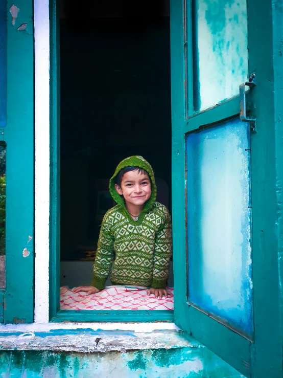 a little girl that is looking out of a window, inspired by Steve McCurry, pexels contest winner, hurufiyya, wearing a green sweater, uttarakhand, blue hoodie, doorway