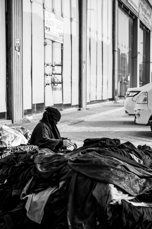 a man sitting on the side of a street next to a pile of garbage, a black and white photo, by Giuseppe Avanzi, beautiful burqa's woman, curled up under the covers, an arab standing watching over, cyberpunk homeless